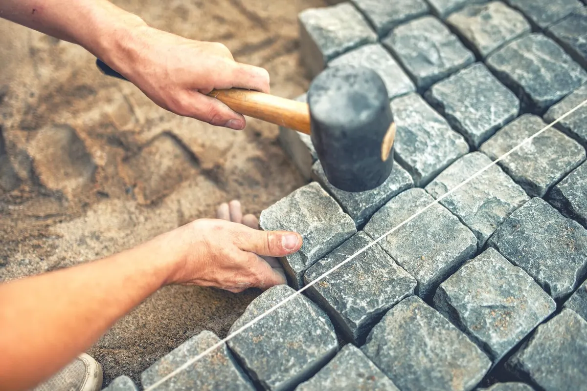 Mains posant des pavés de granit sur un lit de sable avec un maillet en caoutchouc, le long d'un cordeau tendu